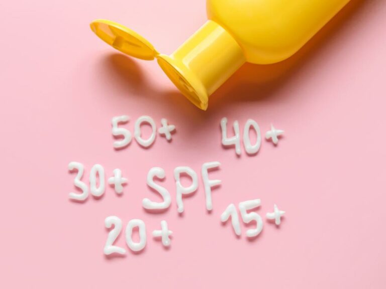 vários números de spf, 15+, 20+, 30+, 40+ e 50+ feitos com protetor solar em uma superfície rosa com um pedaço de garrafa de protetor solar aparecendo