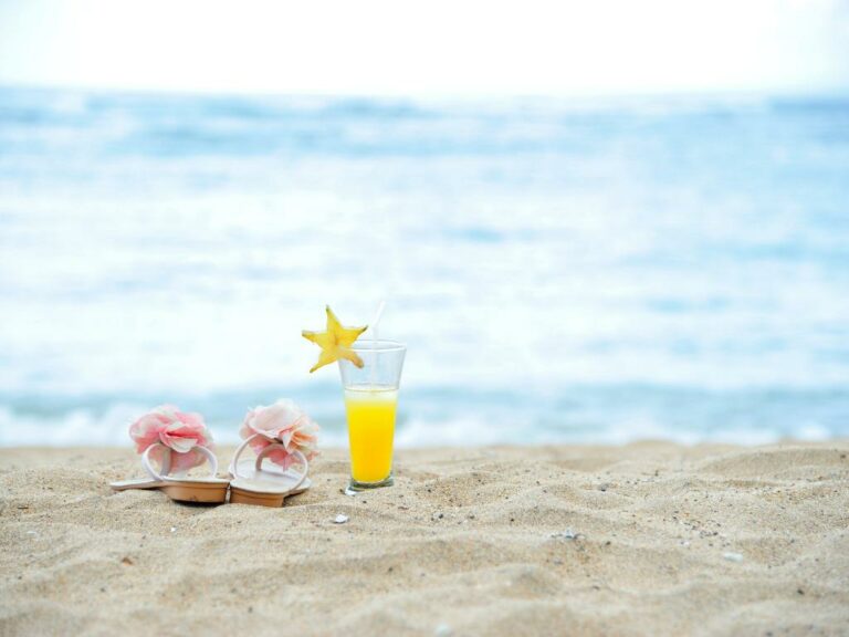 rasterinha bege claro com uma flor cor de rosa em cima das alças com um drink amarelo do lado na areia da praia com o mar de fundo