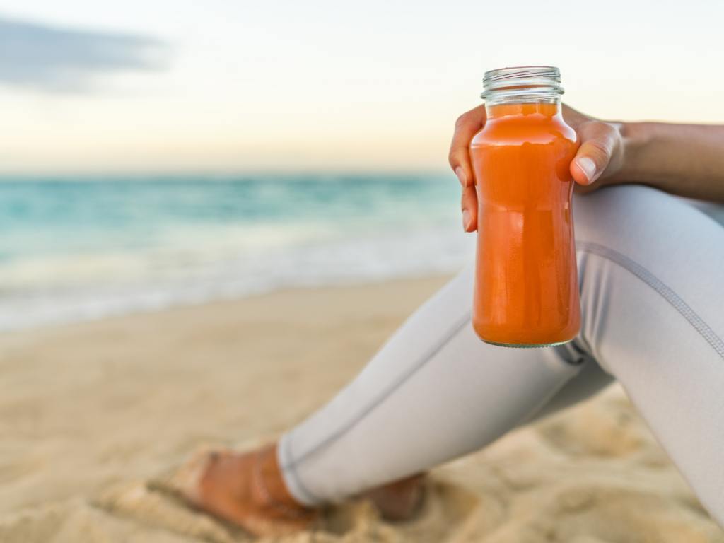 mulher na praia segurando uma garrafa com suco de cenoura sentada na areia