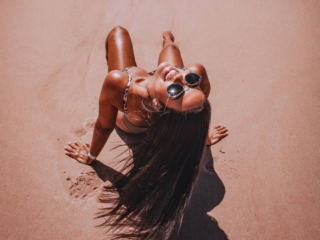 mulher de cabelos longos e oculos escuros com pele bronzeada sentada na areia e sorrindo